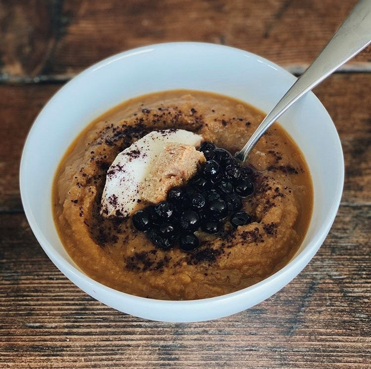 Bowl of orange pureed soup topped with pickled blueberries, blueberry powder, crouton, and cheese