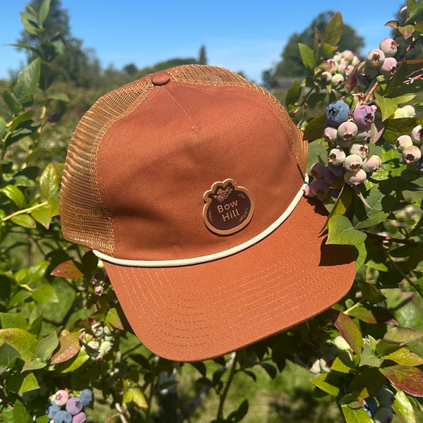 Brown cap with mesh back and Bow Hill logo patch on a blueberry bush with berries and green leaves