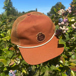 Brown cap with mesh back and Bow Hill logo patch on a blueberry bush with berries and green leaves