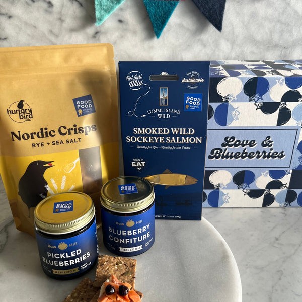 Assorted food products including a bag of Nordic Crisps, smoked wild sockeye salmon, and blueberry confiture on a marble surface.