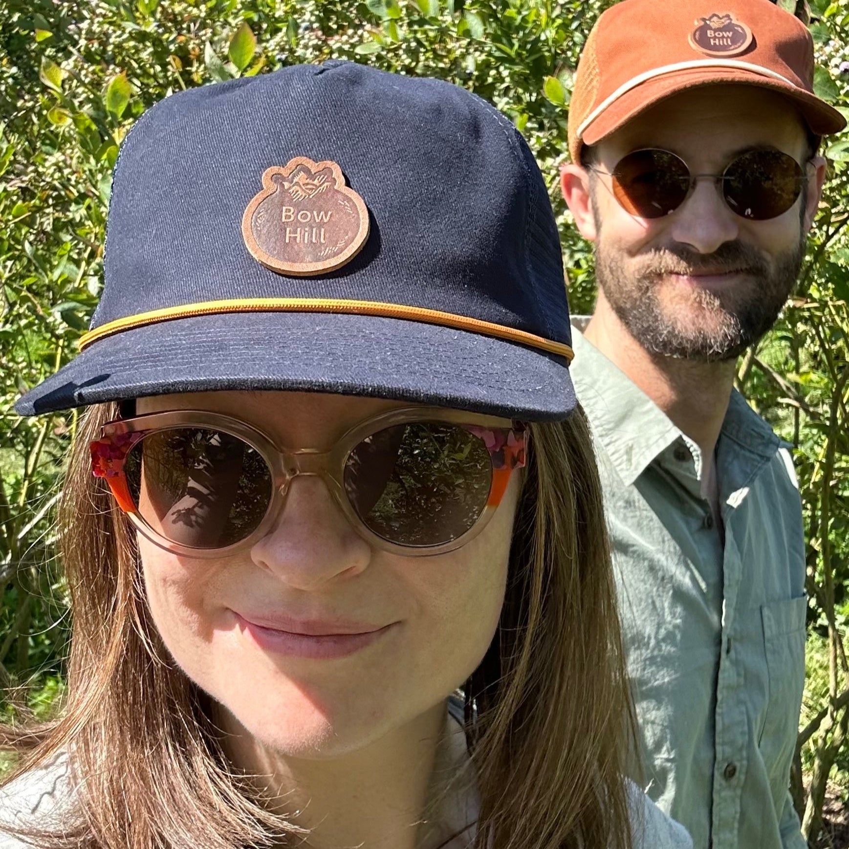 Two people wearing hats and sunglasses outdoors with blueberry bushes in the background